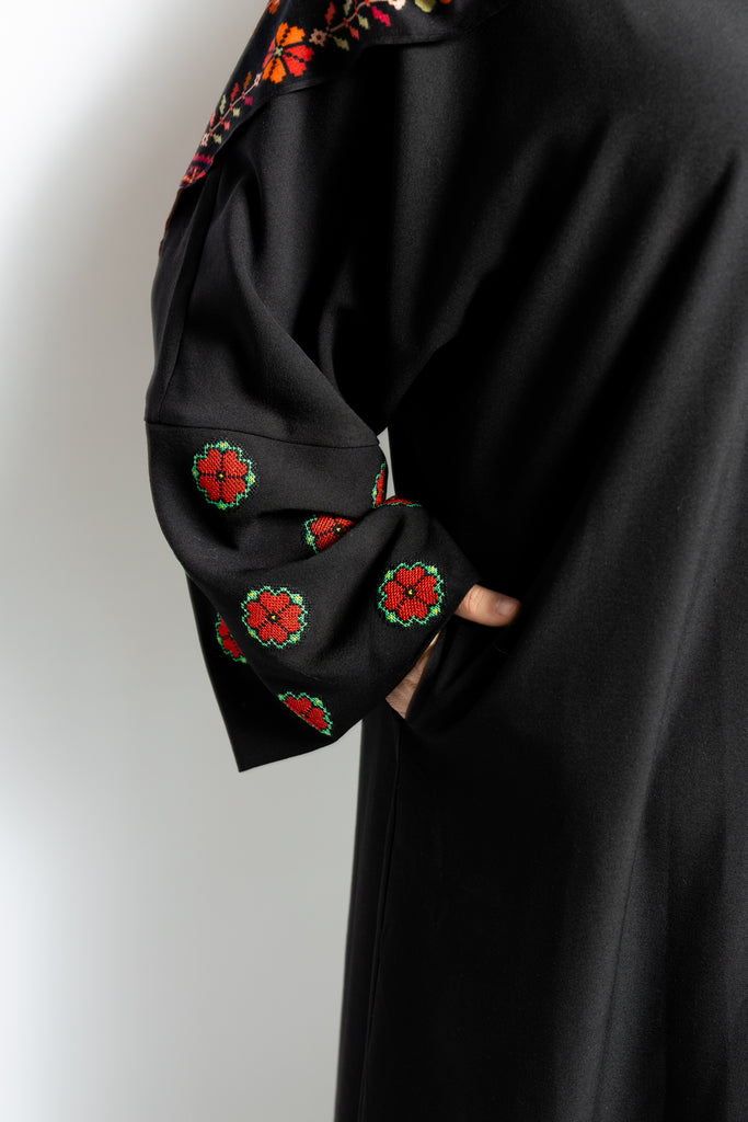 a black abaya with button clasps and pockets and watermelon and poppy tatreez palestinian embroidery along the sleeves 