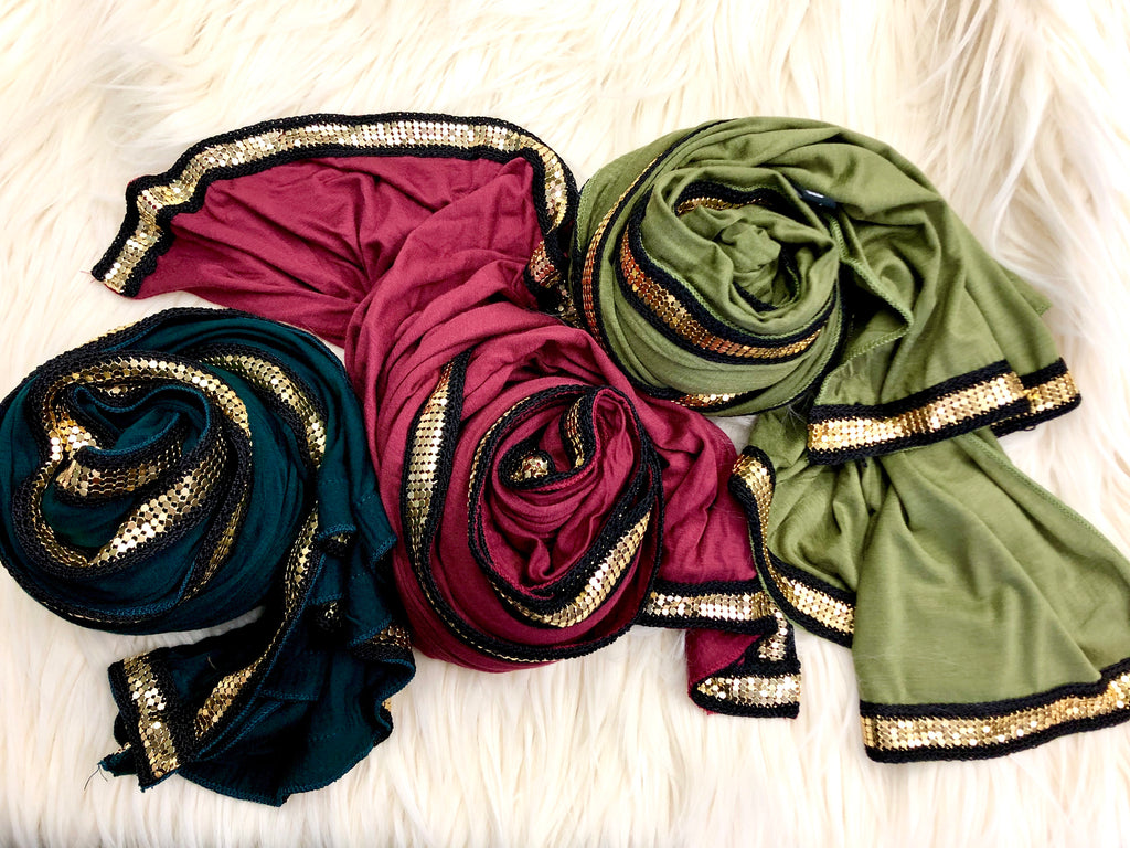 three jersey hijabs embellished with a gold metal trim in maroon, olive, and forest green laid out on a fur rug