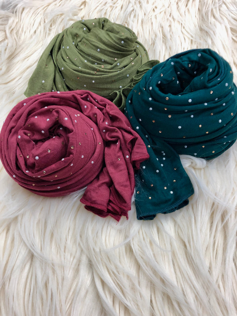 three jersey hijabs embellished with beaded pearls in maroon, olive, and forest green laid out on a fur rug