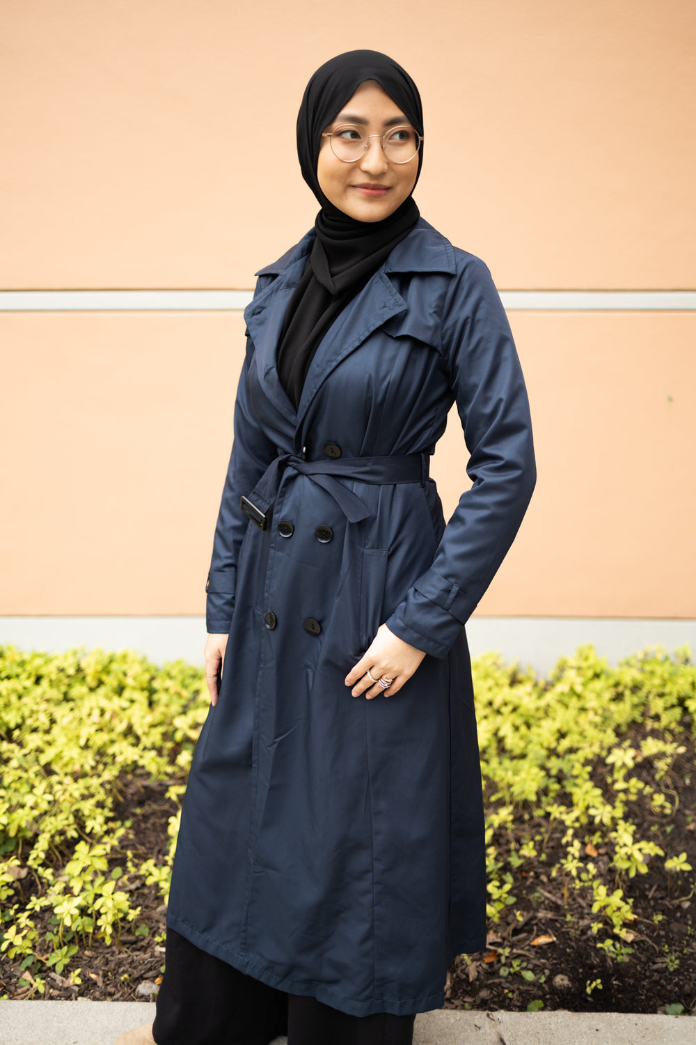 asian muslim woman wearing a black hijab and outfit with a navy blue maxi trench coat with black buttons