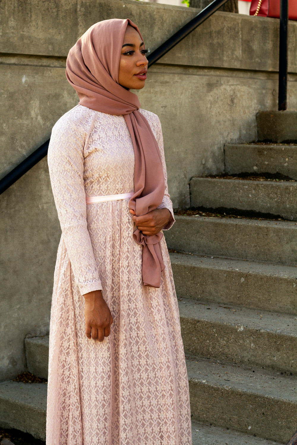 black muslim woman wearing a long sleeve maxi dress in pink lace with a satin waist tie and mauve hijab