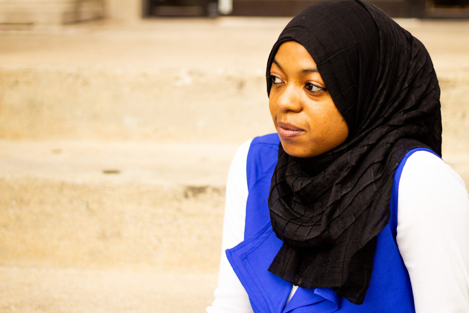black muslim woman wearing a royal blue vest and long sleeved white top with a viscose pleated grid black hijab