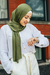 muslim woman checking her watch wearing white top and pants with a beaded olive green hijab