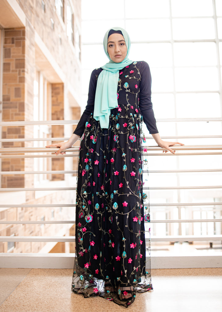hijabi woman wearing a teal chiffon hijab and black long sleeved maxi dress with embroidered florals in pink blue and green