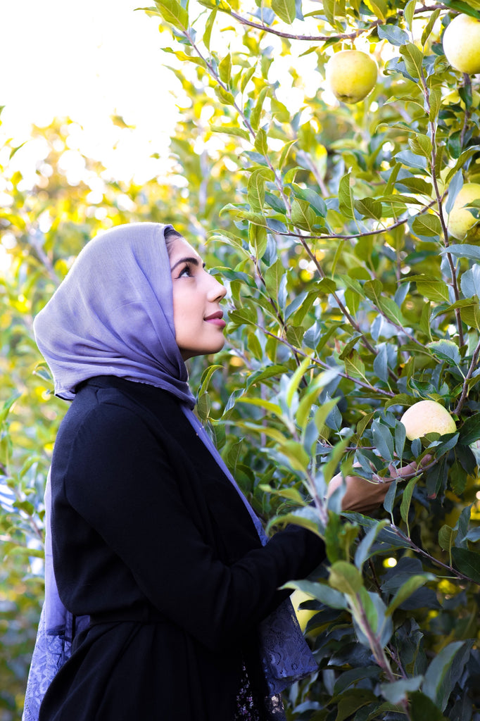 hijabi woman wearing a periwinkle purple modal hijab with lace ends wearing all black in an orchard