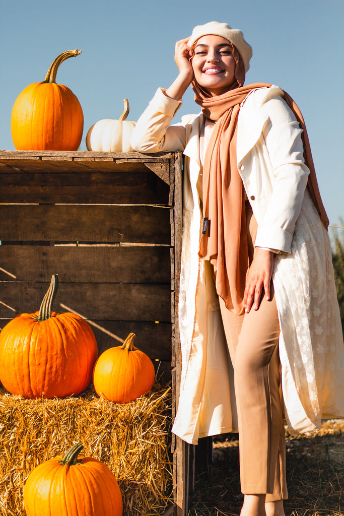 hijabi woman wearing a taupe chiffon hijab surrounded by a pumpkin hay fall theme and wearing a lace jacket