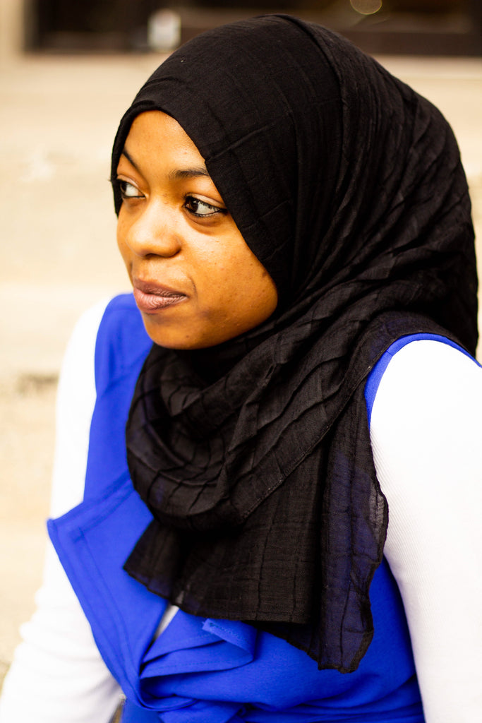 black muslim woman wearing a royal blue vest and long sleeved white top with a viscose pleated grid black hijab
