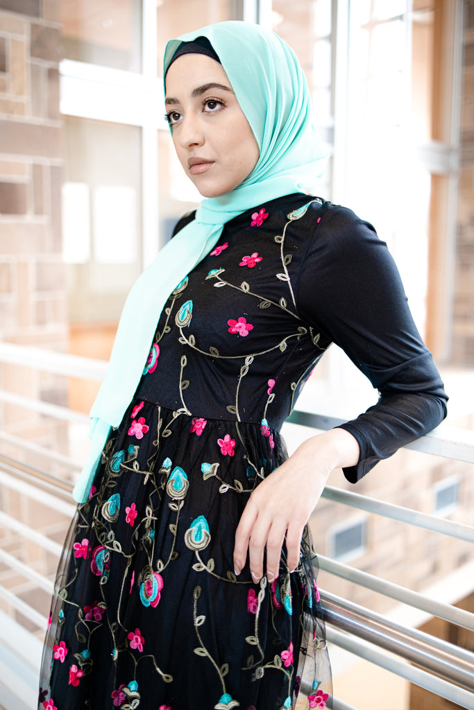 hijabi woman wearing a teal chiffon hijab and black long sleeved maxi dress with embroidered florals in pink blue and green