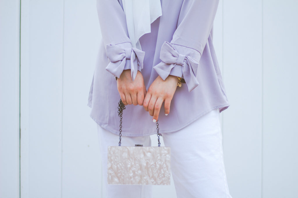 muslim woman wearing a white chiffon hijab and a lilac top with bow sleeves white pants holding a marble purse
